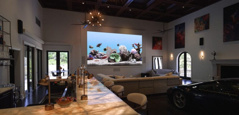 Panoramic view of an ultra-luxury entertainment room in Vero Beach featuring a marble bar, large seating area with projector, and exotic car display under a custom dark wood coffered ceiling by Island Harbor Custom Builders.