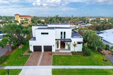 Front aerial view of a modern two-story custom home in Vero Beach featuring white stucco walls, dark flat roof accents, and a minimalist landscape design by Island Harbor Custom Builders.