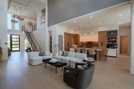 A sprawling luxury open-concept living room in Vero Beach featuring a modern floating staircase, double-height ceilings, a gourmet kitchen with wood grain cabinetry, and light oak floors by Island Harbor Custom Builders.