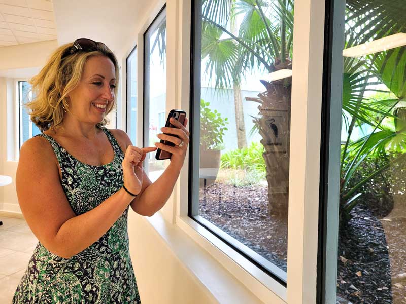 A happy homeowner in Vero Beach using her smartphone in front of newly installed, high-end replacement windows by Island Harbor Custom Builders.