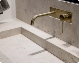 Brushed brass wall-mounted bathroom faucet above a natural stone sink with coordinated limestone finishes.